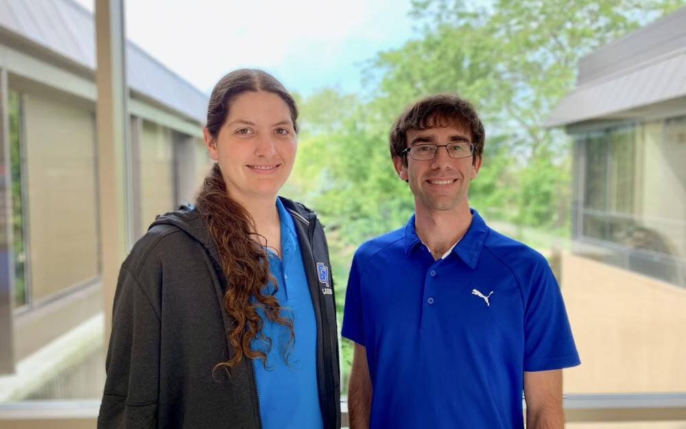 A woman and a man are standing side by side indoors, smiling at the camera with large windows and trees in the background. The woman has long curly hair, wears a blue shirt with a dark jacket, and the man wears glasses and a blue Puma polo shirt.